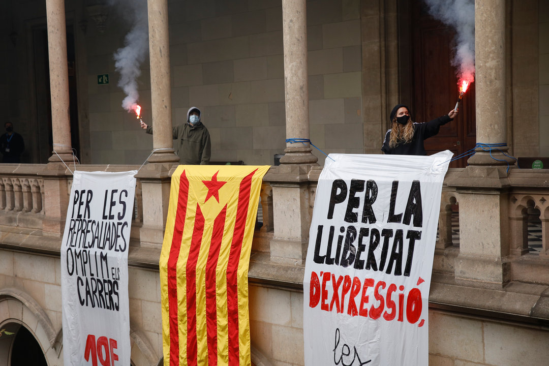 Dos personas sostienen bengalas junto a dos pancartas y una bandera estelada, durante una manifestación contra el encarcelamiento de Pablo Hasel, en el edificio histórico de la Universitat de Barcelona (UB)