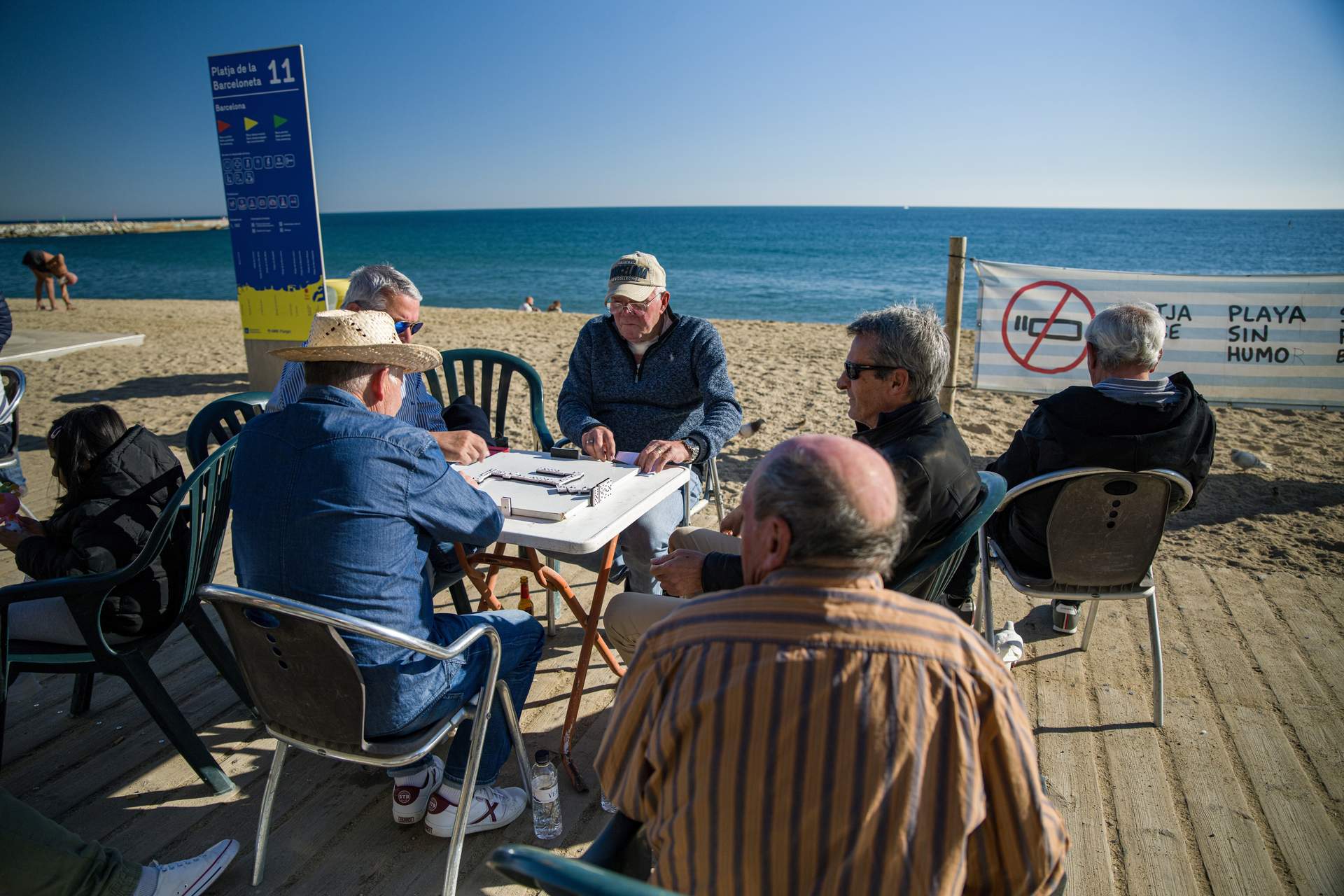 Unos hombres juegan al dominó en la playa de la Barceloneta, al fondo, un cartel de "Playa sin humo". Foto: Lorena Sopêna / Europa Press